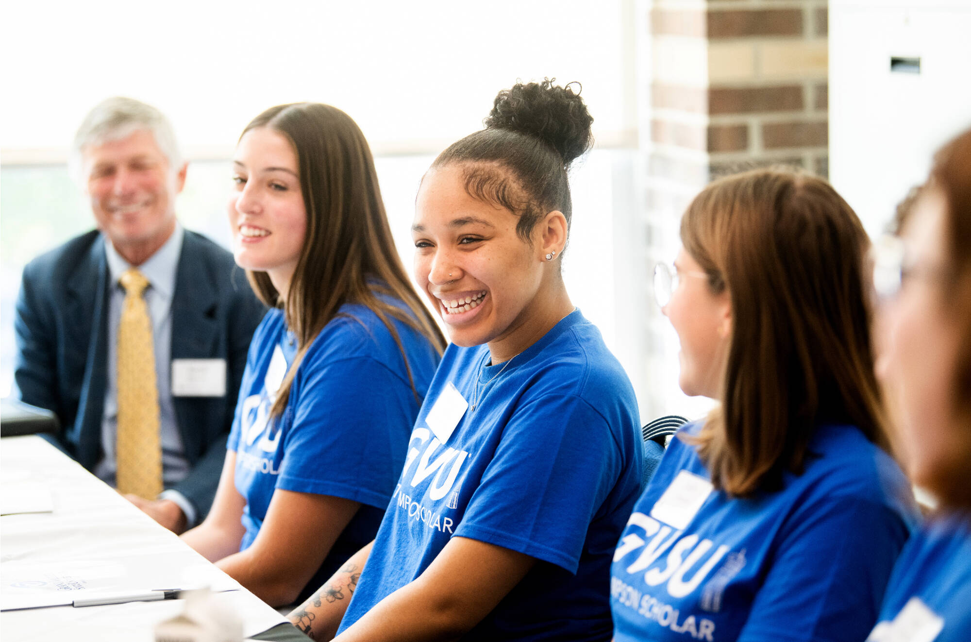 Students wearing blue Thompson Scholar t-shirts smiling with sunlight pouring in from the window behind them.